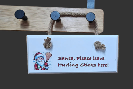 Wooden plaque with Santa Claus illustration and text 'Santa, Please leave Hurling Sticks here!' on a dark background.